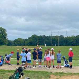 Junior golf group class at Reston National Golf Course driving range, with juniors watching the instructor demonstrate a golf swing.